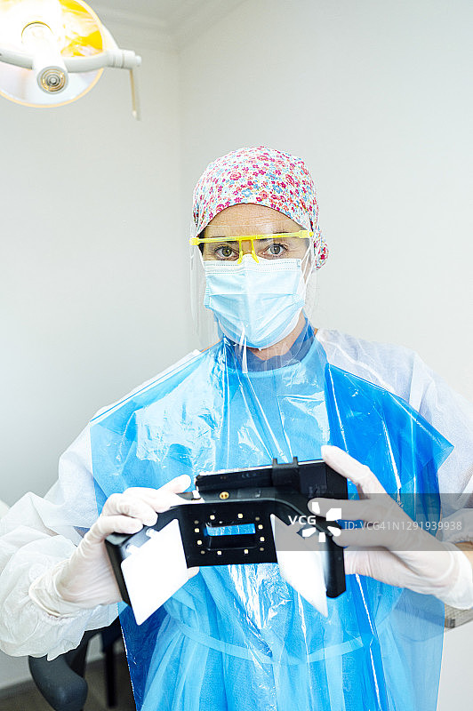 2019冠状病毒病(Covid-19)期间，医生穿着防护工作服，手持移动牙科摄影设备站在诊所图片素材