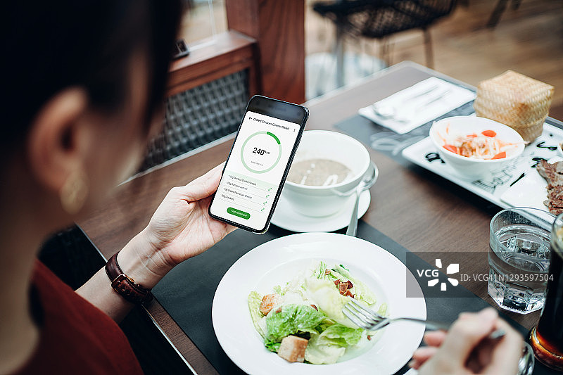 一名年轻的亚洲女性正在智能手机上使用健身计划手机应用来制定自己的日常饮食计划，检查饮食中的营养成分和卡路里摄入量，在餐厅里吃新鲜的凯撒沙拉和蘑菇汤图片素材