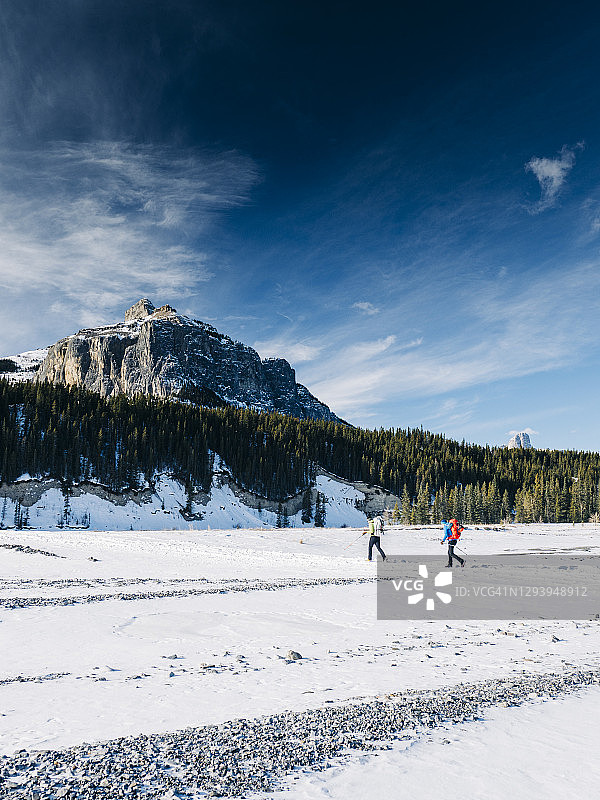 在加拿大落基山脉的东侧，两名冰上登山者背着巨大的背包徒步穿越幽灵河冰冻的河床图片素材