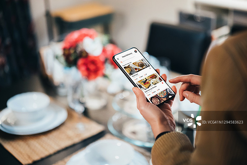 在一个舒适的家里，年轻的亚洲女性在餐桌旁的餐厅用智能手机上的移动应用程序使用送餐服务和在线订餐图片素材