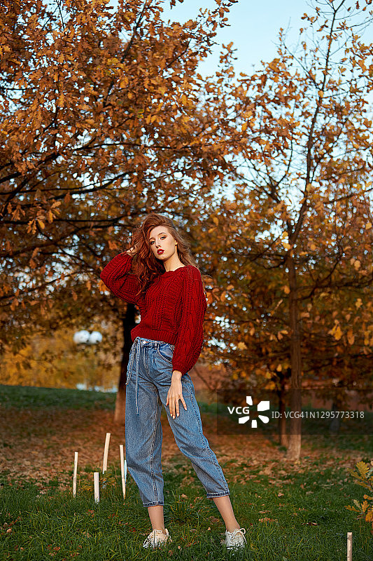 穿着红毛衣的红头发年轻女子在公园散步。秋天美丽的肖像，一个时尚的红发女人在日落图片素材