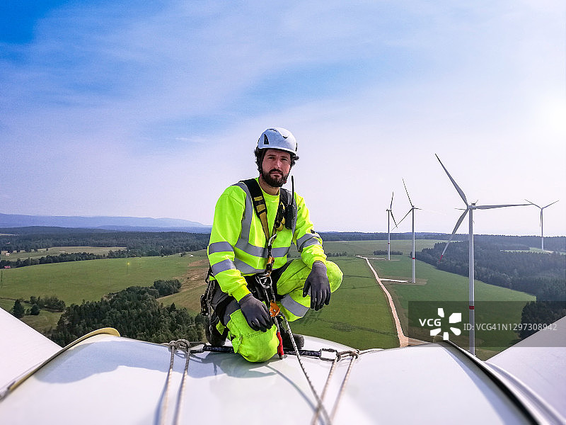 工业攀登者，绳索获取技术人员跪在巨大的陆上风力涡轮机上准备检查叶片身后的风力发电场图片素材