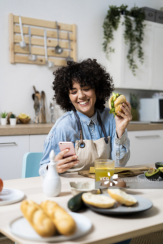 一名年轻女子拿着三明治在厨房的桌子上使用智能手机的肖像图片素材
