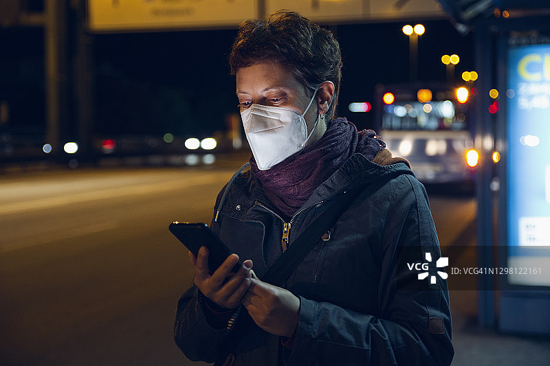 2019冠状病毒病期间，佩戴防护口罩、在公交车站使用智能手机的女性图片素材