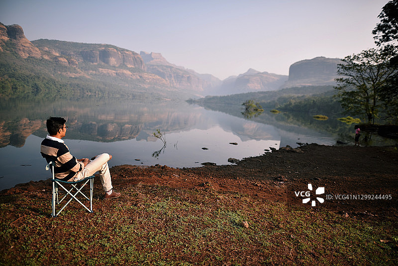 男人坐在椅子上，放松在湖边，树和山图片素材