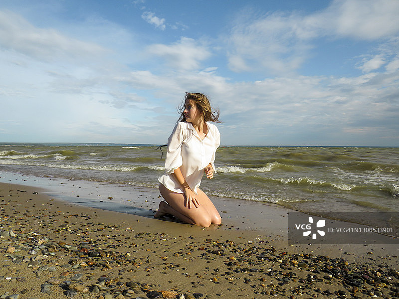 一位头发蓬乱的金发女郎，穿着白色衬衫和比基尼，一双晒黑了的腿修长在沙滩上，享受着海风和阳光的温暖图片素材