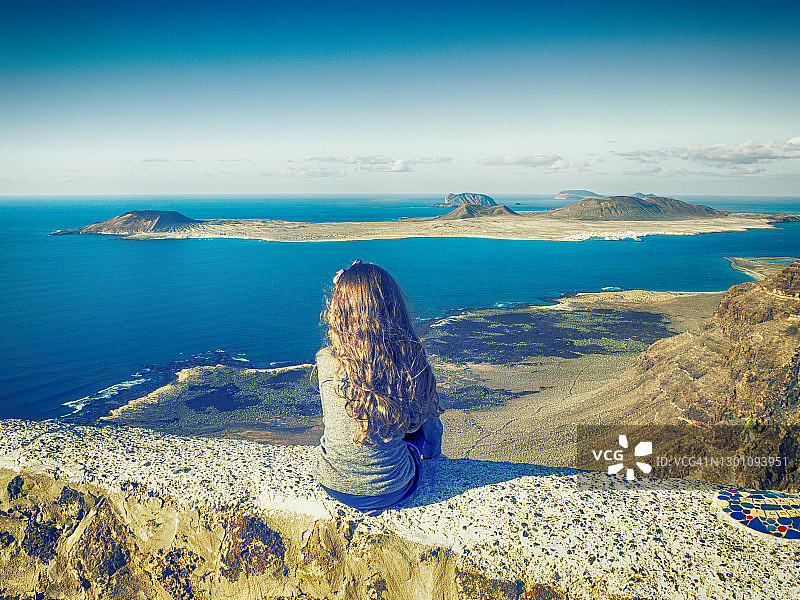 一个女孩坐在山上看风景的后视图，格雷西奥萨，加那利群岛，西班牙图片素材