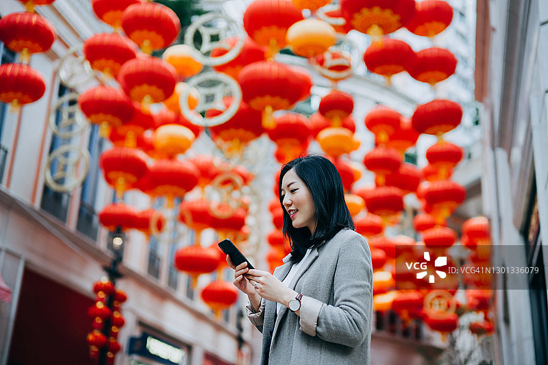微笑的年轻亚洲女子在城市里使用智能手机，街道上挂着传统的中国红灯笼作为背景。中国传统文化，节日庆典主题图片素材
