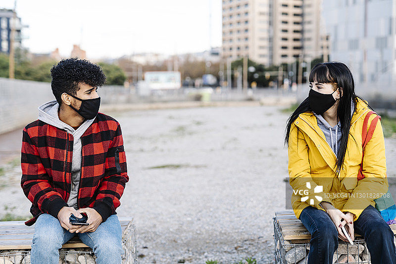 2019冠状病毒病期间，戴着防护口罩坐在社交距离较远的长椅上的夫妇图片素材