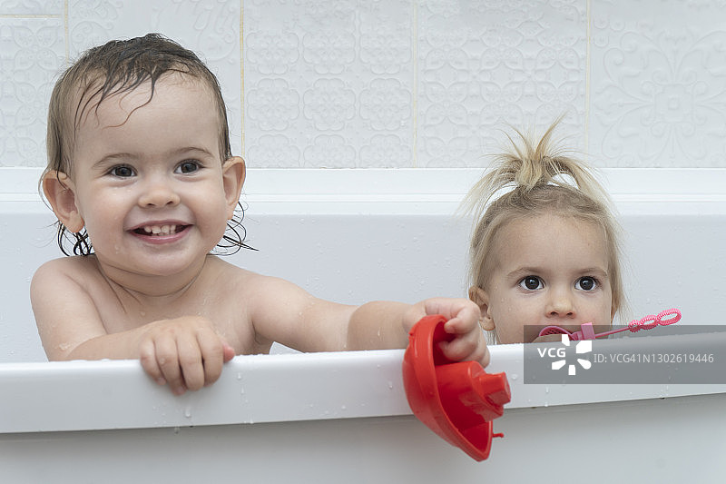 肖像的两个小高加索可爱的女孩姐妹坐在浴缸，有时间洗澡，玩洗澡玩具，微笑，白色的背景，复制空间。图片素材