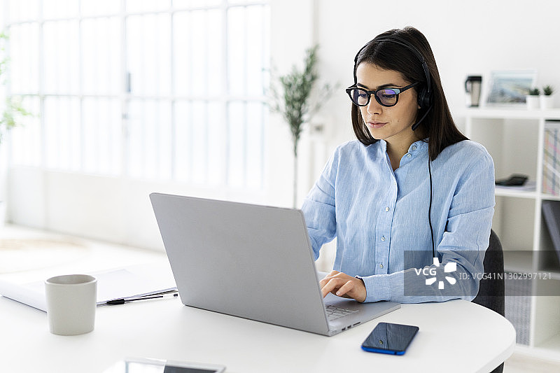 女性客户服务代表，戴着耳机坐在办公室用笔记本电脑工作图片素材