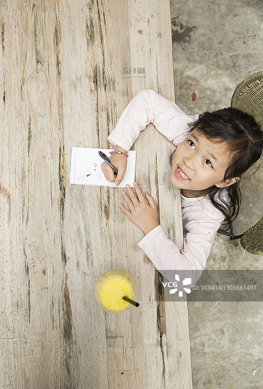 亚洲小女孩看着照相机，在一张木桌上画画。图片素材