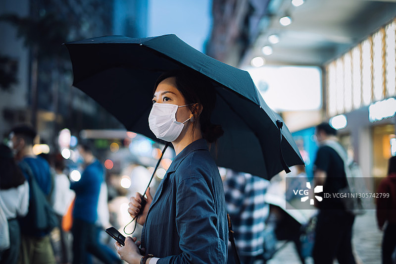 年轻的亚洲妇女戴着防护口罩和雨伞在雨中穿过繁忙的市中心街道，对抗行人，并照亮城市的城市场景在晚上。日常生活的新常态图片素材
