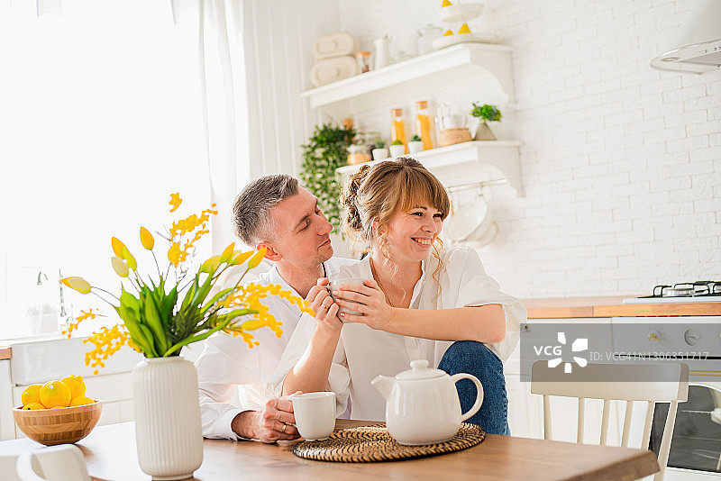 在现代厨房的背景上，一对幸福的年轻夫妇坐在一张桌子旁，手里拿着杯子图片素材