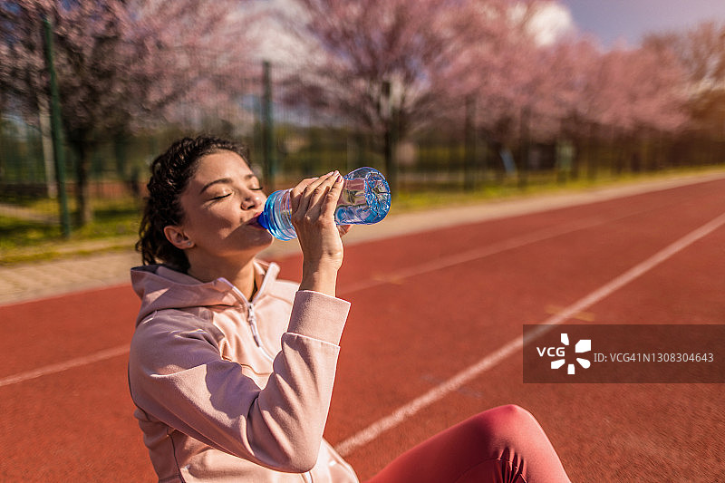 运动的年轻女子在运动轨道上跑步后喝水。图片素材