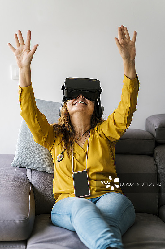 成熟的女人戴着虚拟现实耳机坐在家里的沙发上打手势图片素材