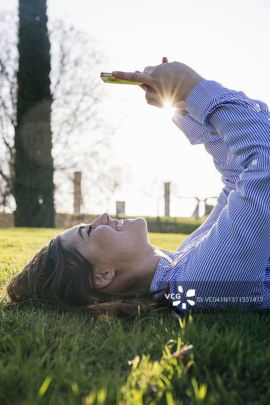 在阳光明媚的日子里，躺在草地上玩手机的快乐女人图片素材