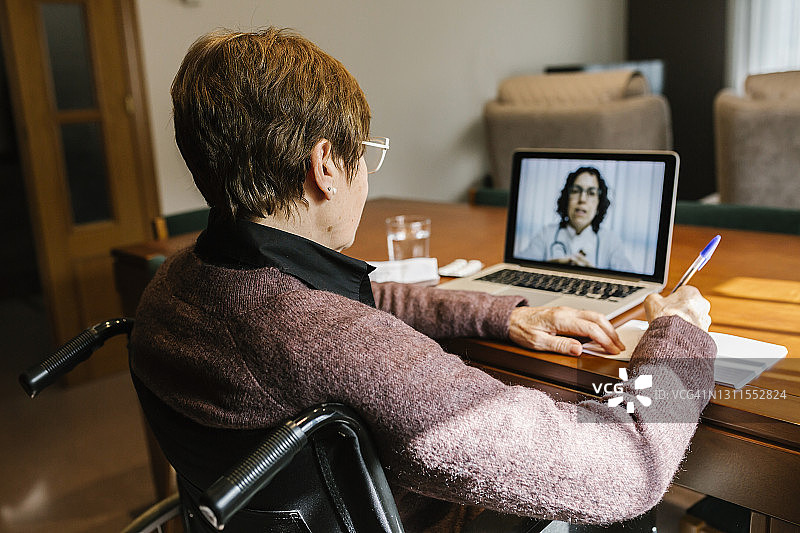 2019冠状病毒病(COVID-19)期间，坐在轮椅上写书、在家视频咨询的老年妇女图片素材