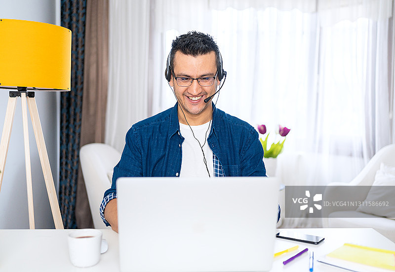 微笑的年轻人使用电脑笔记本电脑和耳机在线会议。在家远程工作图片素材