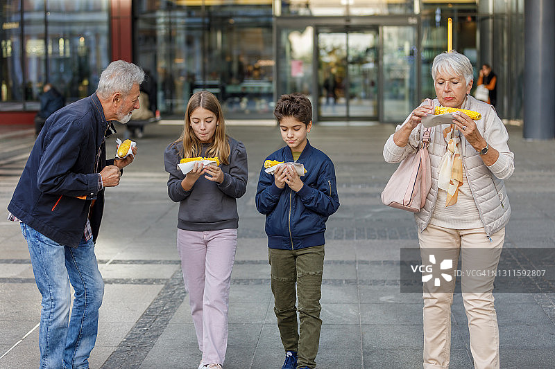 一对老年夫妇和孙辈们在城市街道上吃玉米。图片素材