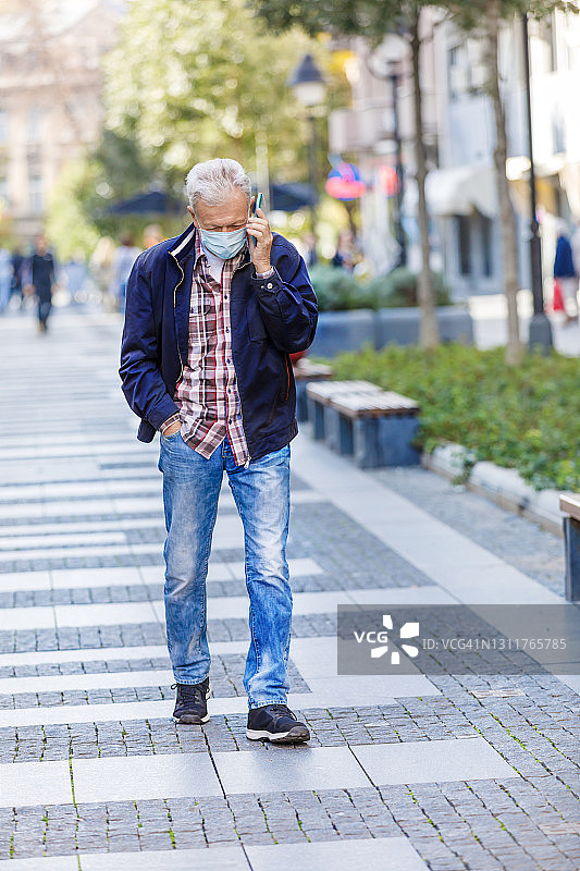 一位老人在流感大流行期间匆忙地使用手机行走在城市街道上。图片素材