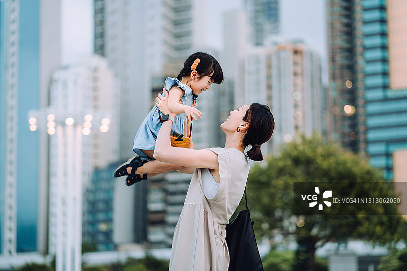 在公园里，一位可爱的年轻亚洲母亲将可爱的小女儿举到空中，映衬着城市的天际线。他们互相看着，笑得很开心。享受亲密的家庭时光图片素材