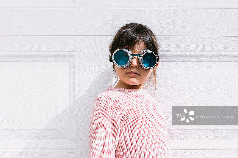 黑头发的小女孩，粉色毛衣，戴着维多利亚时代的眼镜，站在一间车库的白色门口，看着摄像头图片素材