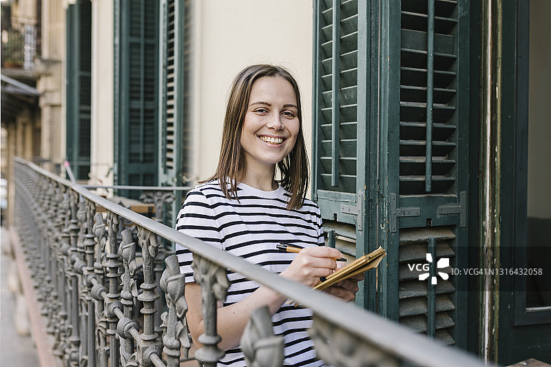 面带微笑的女人拿着笔和日记站在阳台的栏杆旁图片素材