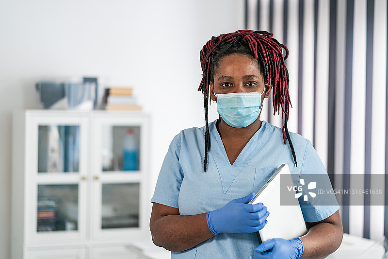 新冠肺炎疫情期间，中年非洲裔女医生手持笔记本电脑，在医生的办公室里戴着防护口罩图片素材
