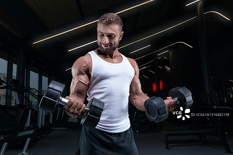 穿着白色t恤的肌肉男拿着哑铃在健身房锻炼。二头肌泵。健身和健美概念。图片素材
