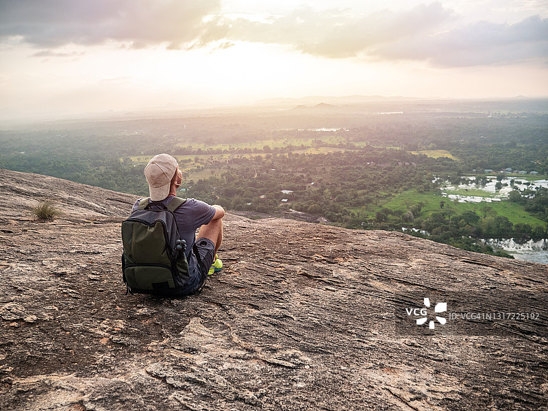 后视图的年轻男子坐在岩石顶部在日落凝视美丽的丛林自然景观图片素材
