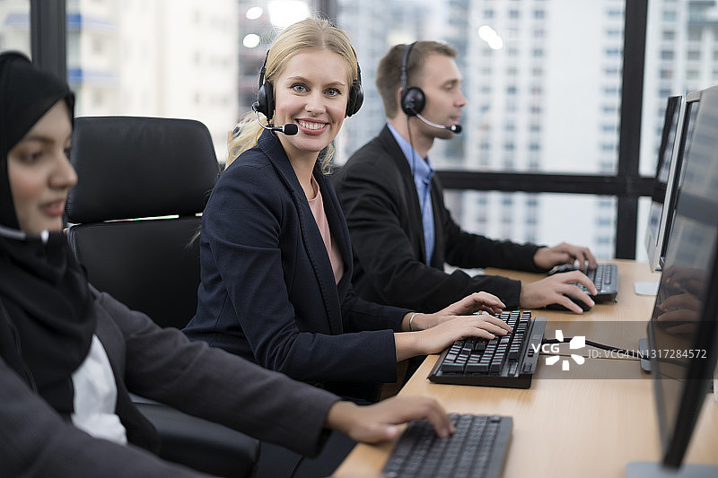 近距离观察微笑的女性呼叫中心员工戴着耳机在工作场所与同事在办公桌上使用电脑。图片素材