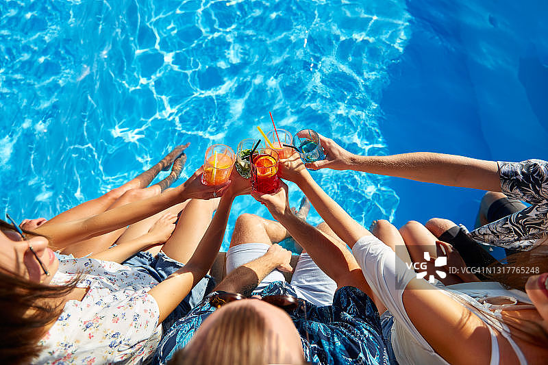 顶视图的朋友碰杯与新鲜多彩的鸡尾酒坐在游泳池在阳光明媚的夏天。在热带度假的豪华别墅泳池边派对上，人们举杯敬酒图片素材
