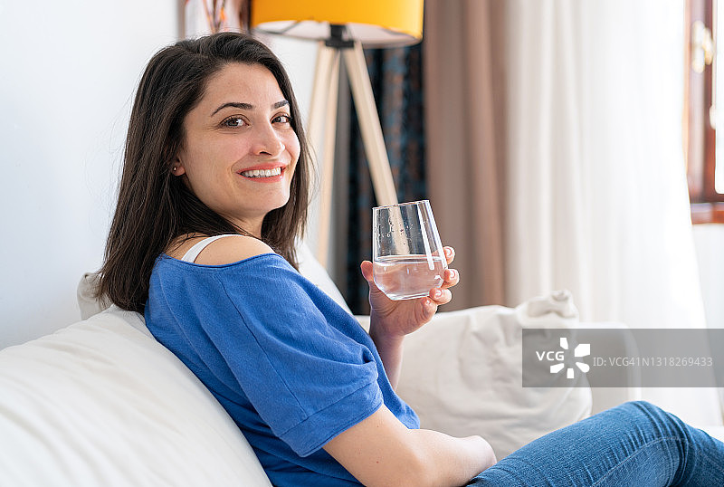幸福的年轻女子微笑着，在家里拿着一杯水。坐在客厅沙发上的女人一边喝着水一边放松微笑。喝足够的水对健康的好处图片素材