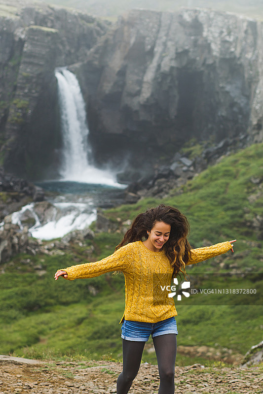 在冰岛，穿着橙色毛衣的快乐卷毛女人笑着和瀑布赛跑。风在头发图片素材