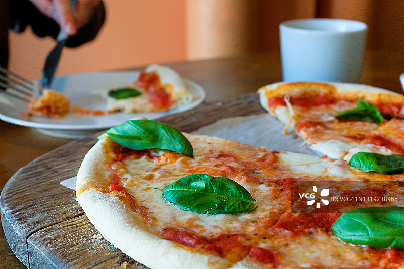 圆形玛格丽塔披萨，配马苏里拉奶酪和罗勒叶，放在木架或砧板上，咖啡馆或快餐店的桌子上。旁边，一个女孩或女人切下一片披萨吃了。传统的意大利美食。素食。图片素材