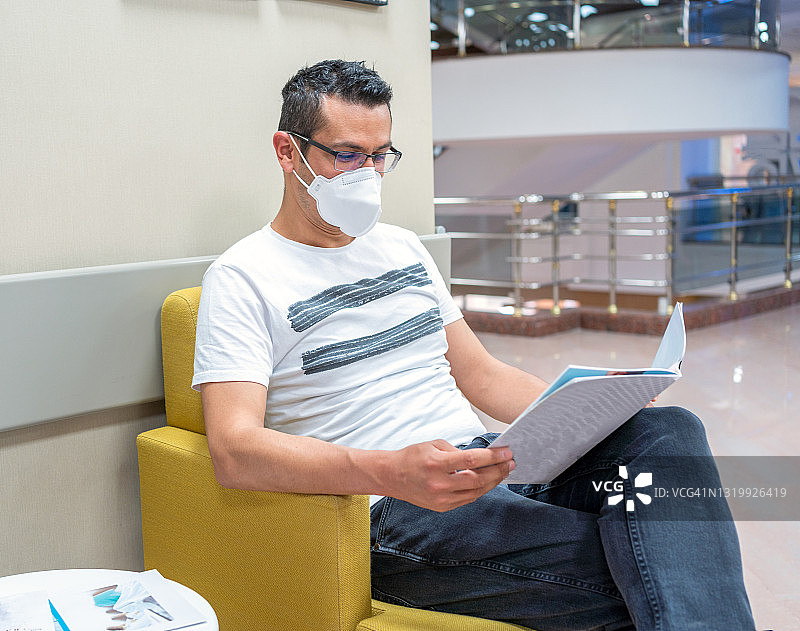 男病人戴着防护口罩坐在等候区，在医院看杂志。体检或身体检查图片素材