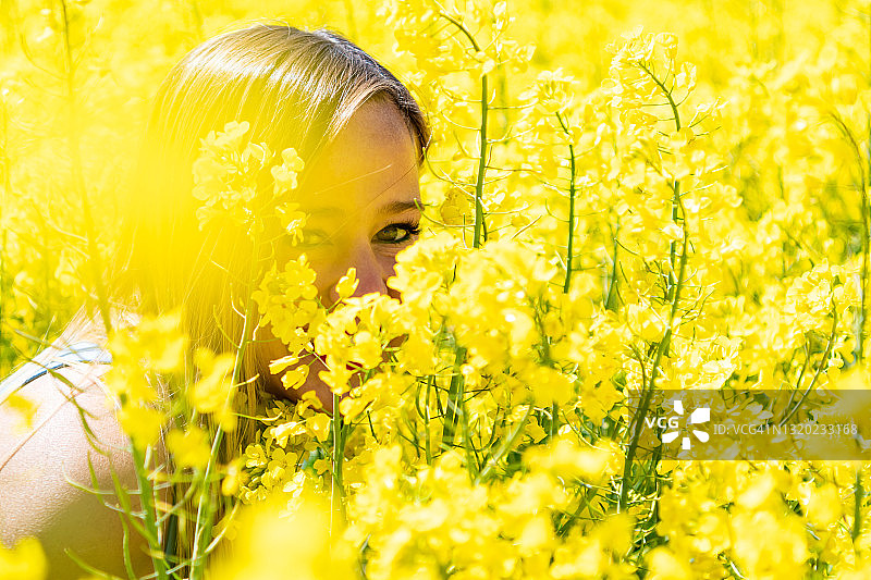 金发女人蹲在许多黄色的花。前景中有几张照片，形成了黄色的滤镜效果。这些花被称为油菜籽或菜籽油。图片素材
