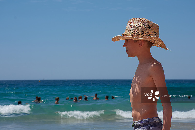 在西班牙贝尼多姆，一个白人孩子穿着泳衣，赤膊，戴着草帽站在海边，看着地中海。图片素材