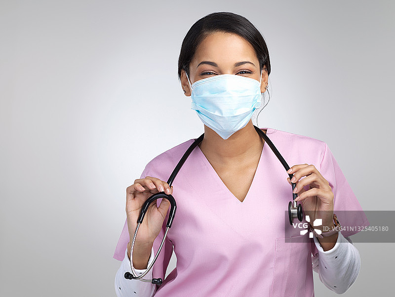 裁剪的肖像，一个迷人的年轻女性保健工作者戴着面具和听诊器，而站在工作室的灰色背景图片素材