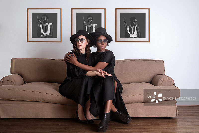 宁静的黑人和白人妇女在黑色连衣裙和软呢帽上的沙发图片素材