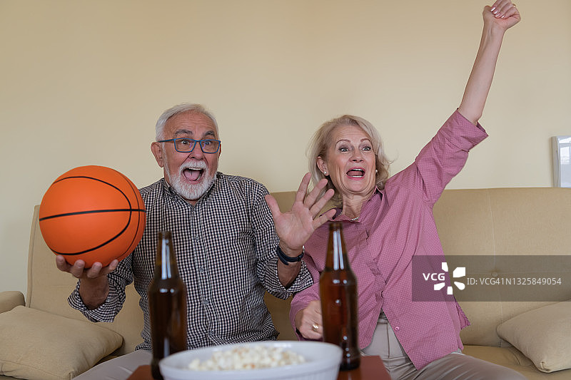 一对快乐的老年夫妇正在家里看篮球比赛。图片素材