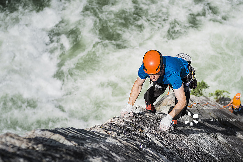 一名男性攀岩者在攀登高耸于汹涌河流之上的戏剧性岩壁时，伸手去抓一只手图片素材