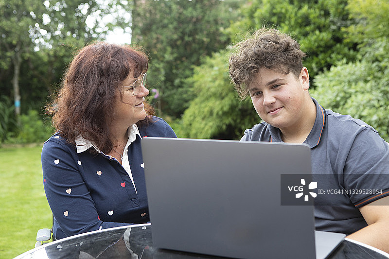 一个十几岁的男孩和他的母亲坐在花园的桌子旁，两人都在看笔记本电脑图片素材