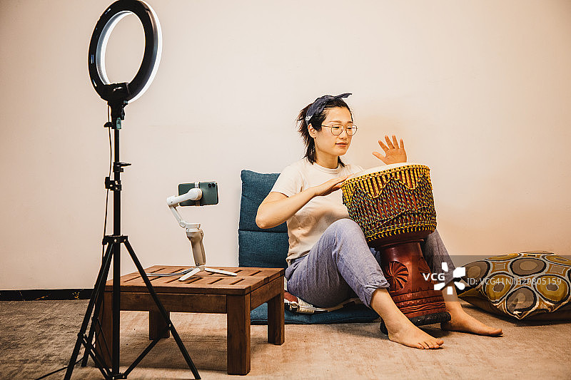 亚洲女人在三脚架上通过手机直播的同时演奏djembe图片素材