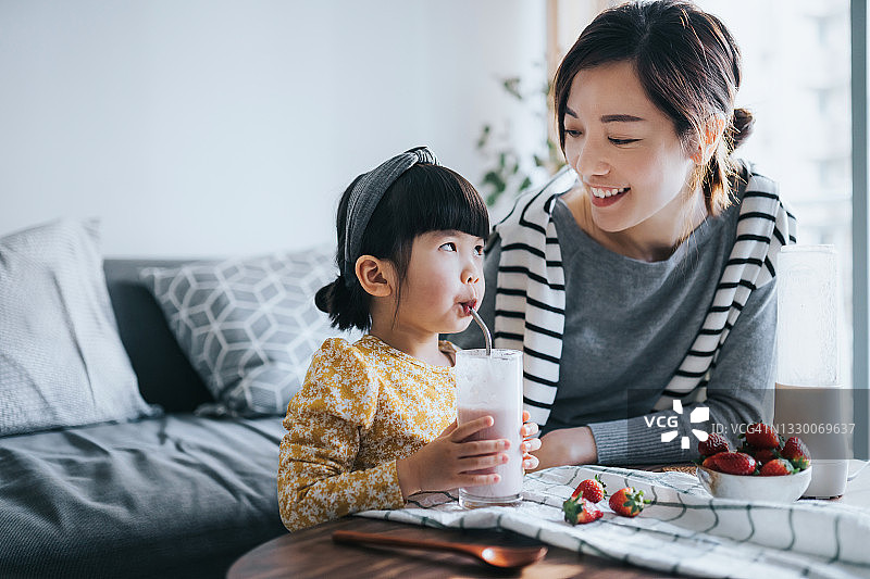 年轻的亚洲母亲和可爱的小女儿在家做健康的自制草莓奶昔。母女相视而笑，享受着饮料。健康的饮食和生活方式图片素材