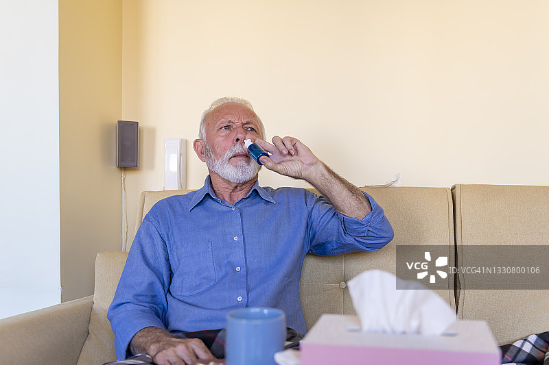 一位患有鼻炎的老人坐在家里的沙发上使用鼻喷雾剂。图片素材