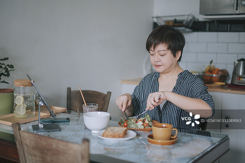 亚洲华人女性在餐桌上一边吃健康的沙拉早餐，一边用数字平板电脑听音乐广播图片素材
