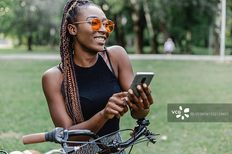 年轻女子站在公园里，手里拿着手机，倚在自行车上图片素材