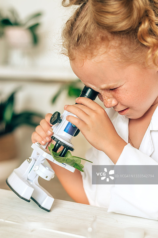 小女孩穿着白色制服，戴着防护眼镜，在一个家庭实验室里看着显微镜。回到学校的概念。年轻的科学家。自然科学。儿童的学前和学校教育图片素材